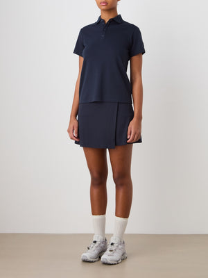 A person stands against a plain background wearing a We Norwegians Medalist Piqué Polo Women in navy, a matching tennis skirt, white socks, and gray athletic sneakers. Their face is not visible.
