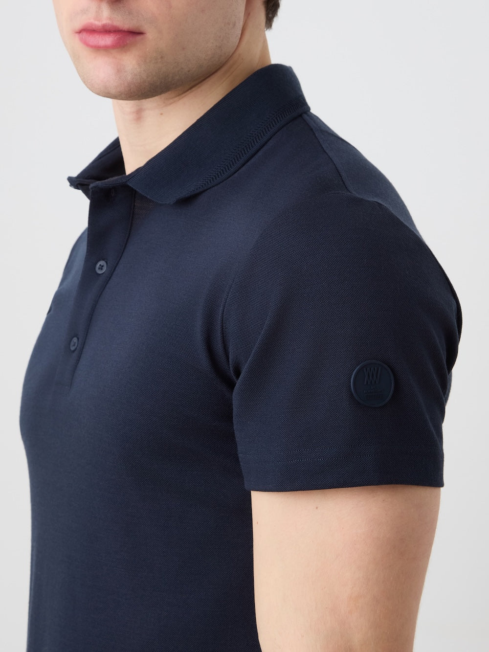 A man wears the We Norwegians Medalist Piqué Polo Men in dark navy; only his upper torso and part of his face are visible. A circular logo patch accents the sleeve, lending this polo shirt a refined touch.