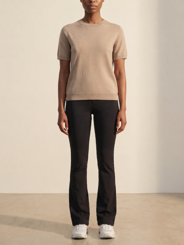 A person stands indoors against a neutral background, wearing the We Norwegians Club Top Women - Summer Cashmere in beige, paired with black pants and white sneakers. Arms are relaxed at their sides; face is not visible.