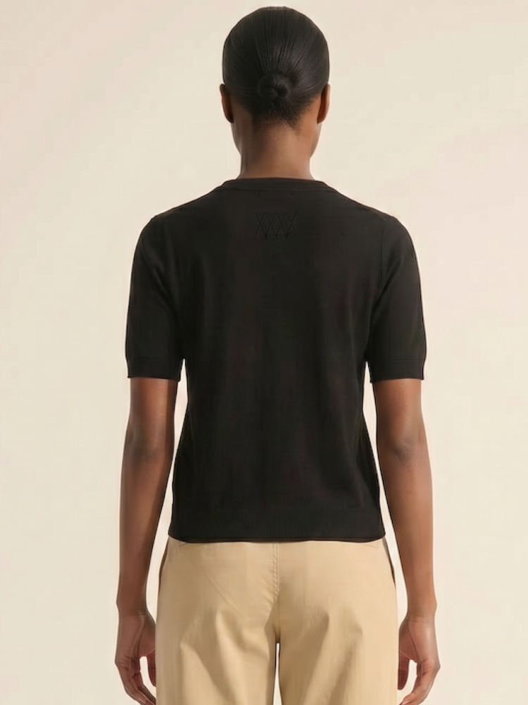 A person with a low bun faces away from the camera, wearing the We Norwegians Club Top Women - Summer Cashmere in black and beige pants against a plain, light background.