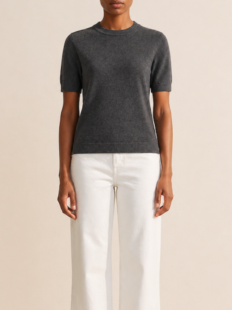 A person stands against a neutral background wearing the We Norwegians Club Top Women - Summer Cashmere in dark gray with high-waisted, wide-leg white pants, arms relaxed by their sides.