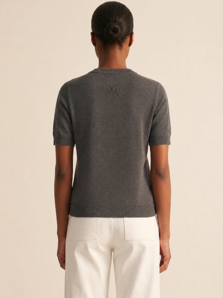A person with a low bun faces away, wearing the We Norwegians Club Top Women - Summer Cashmere in dark gray, featuring subtle stitched details, paired with high-waisted white pants against a plain neutral background.