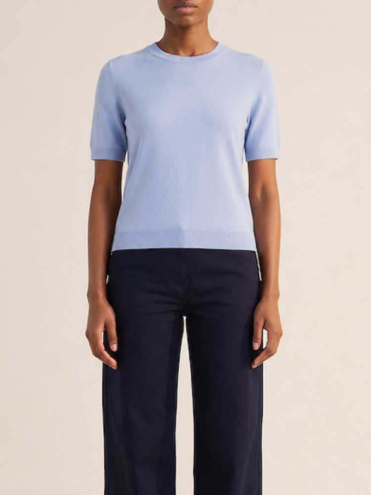 A person stands facing forward against a neutral background, wearing We Norwegians’ Club Top Women - Summer Cashmere in light blue with dark navy blue pants. Their arms are relaxed at their sides.