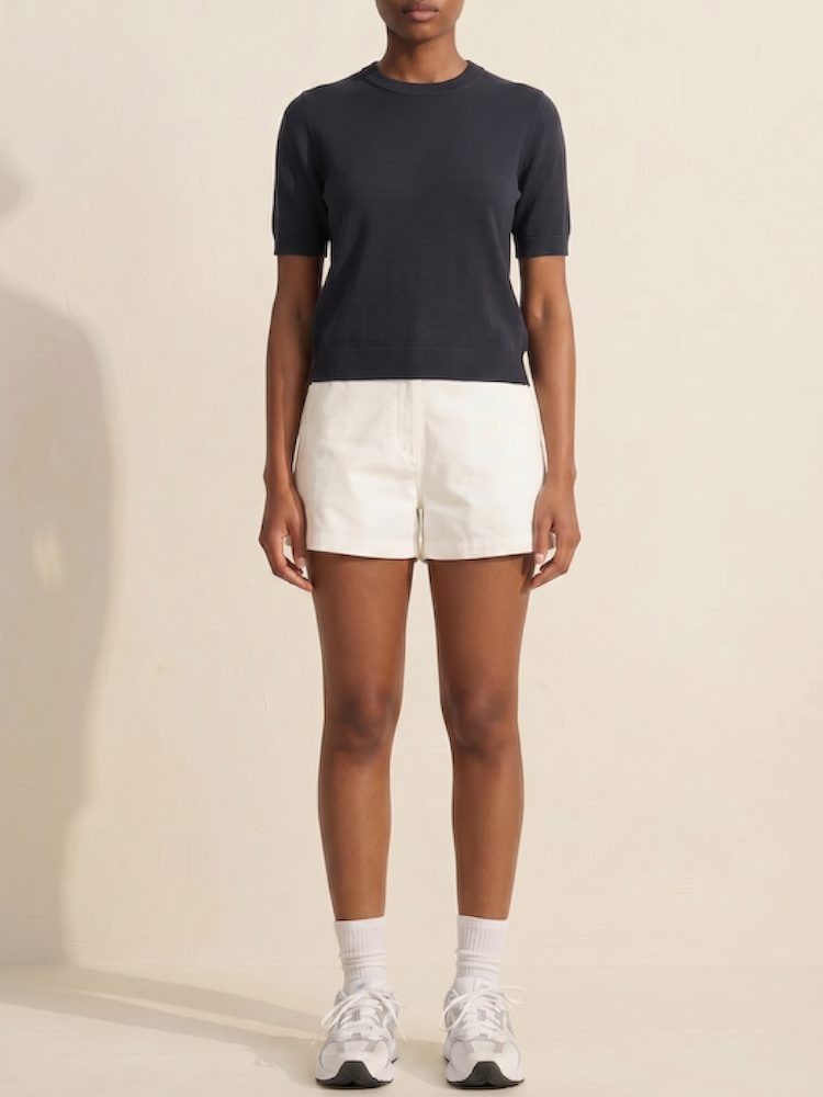 A person stands against a plain light background, wearing the We Norwegians Club Top Women - Summer Cashmere in black, paired with white shorts, crew socks, and athletic sneakers. Their arms are relaxed at their sides.