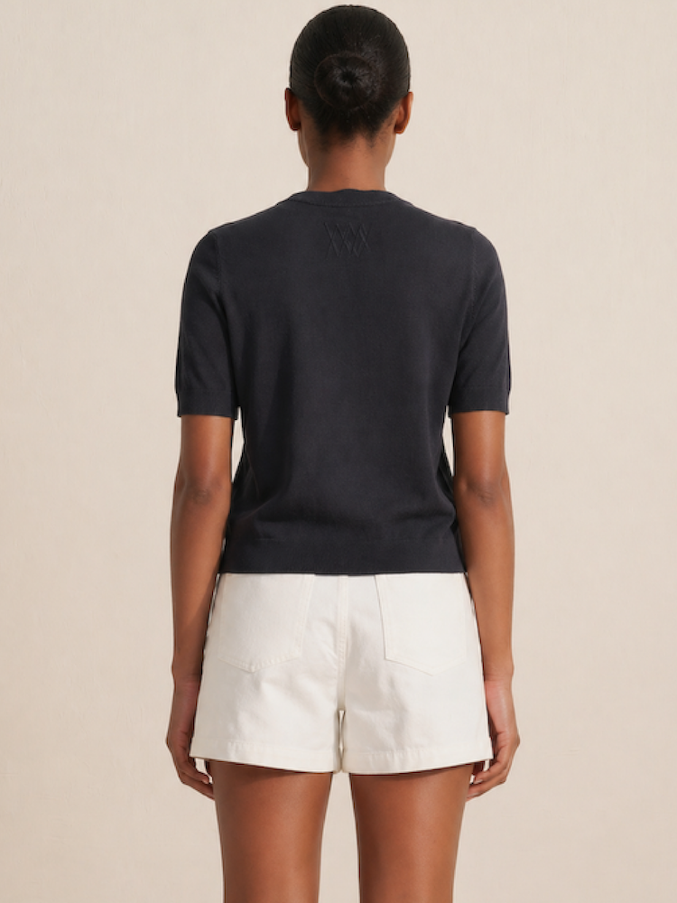 A person faces away, wearing the We Norwegians Club Top Women - Summer Cashmere in dark color with white shorts. Their hair is styled in a neat bun against a plain, neutral background.