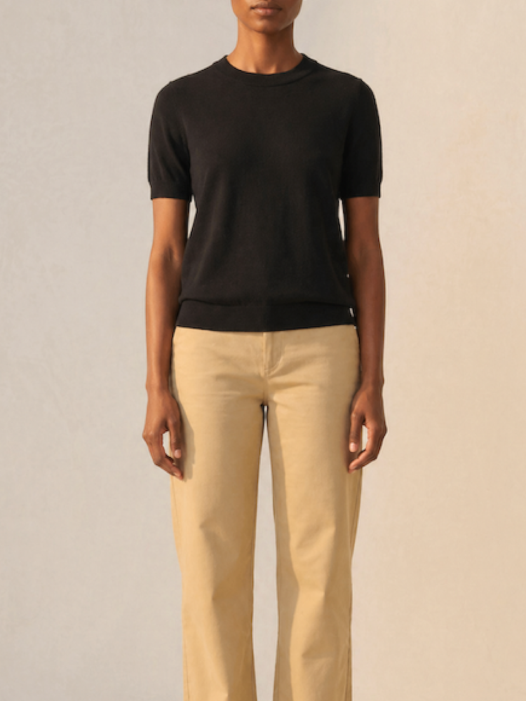 A person stands against a plain light background, wearing the We Norwegians Club Top Women - Summer Cashmere in black and beige pants. Their face is not fully visible.