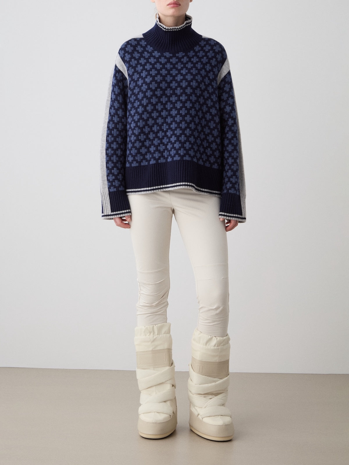 A person stands indoors against a plain background, wearing the We Norwegians Boat Sweater Women in navy, paired with off-white fitted pants and padded snow boots. Their head is slightly turned with hands resting by their sides.