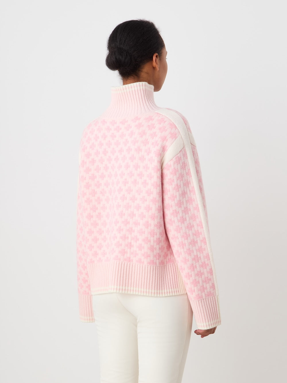 A person faces away, wearing We Norwegians' Boat Sweater women - Pre Spring 26, an oversized light pink and white patterned merino-cashmere turtleneck with long sleeves and cream pants, set against a plain white background.