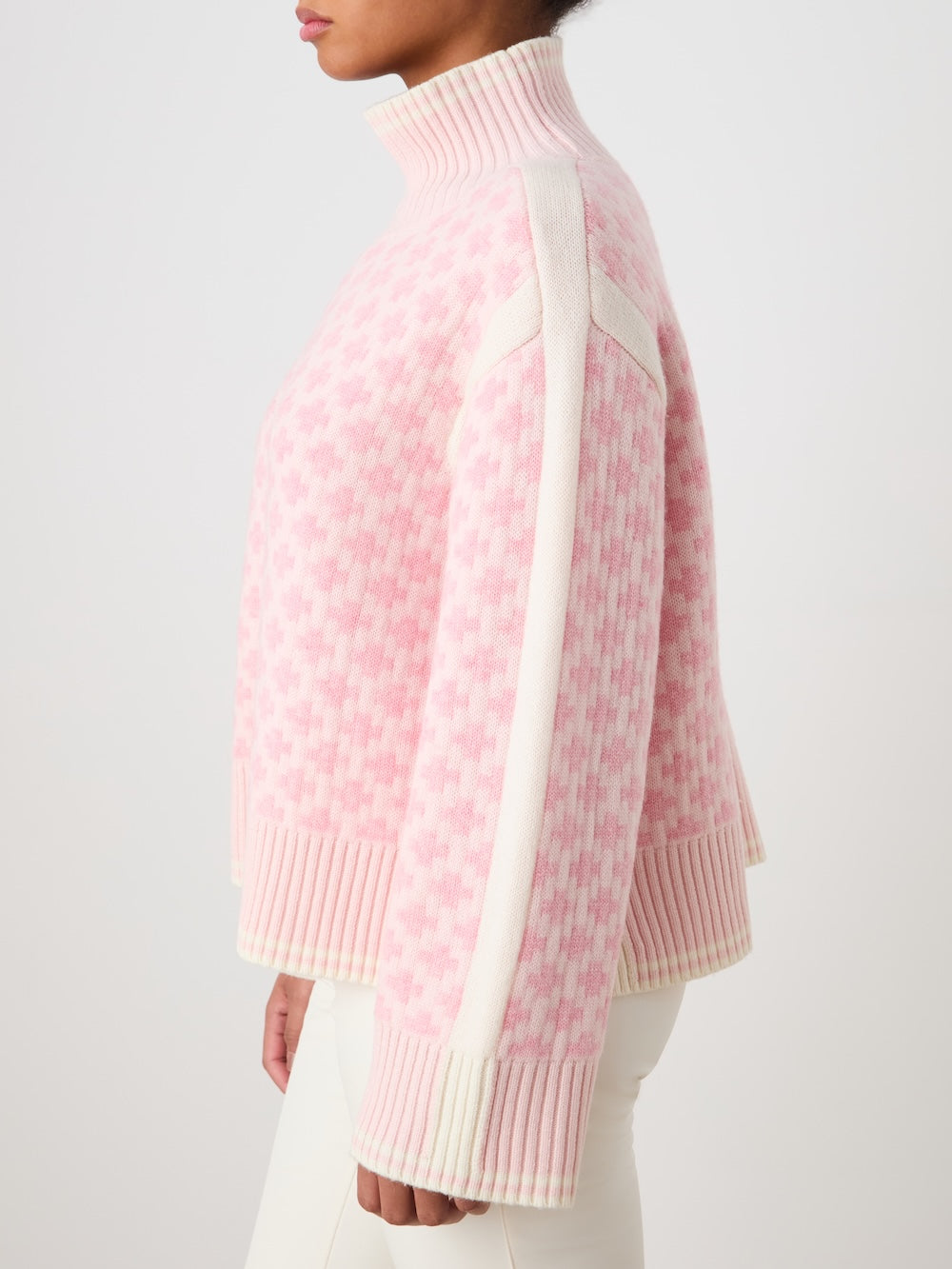 A person wears the We Norwegians Boat Sweater women - Pre Spring 26, an oversized light pink and white patterned knit turtleneck with ribbed cuffs and hem, paired with white pants against a plain light background.