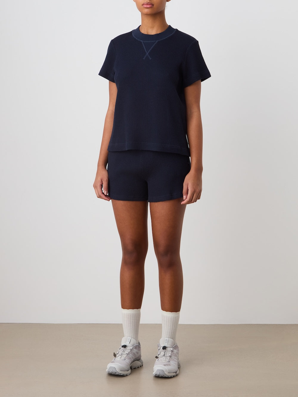 A person stands against a plain background, wearing We Norwegians Waffle Shorts for women, paired with a navy blue short-sleeved top, white crew socks, and grey athletic sneakers. Their arms hang relaxed by their sides.