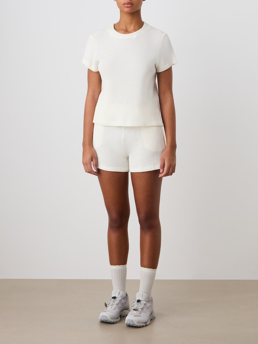 A woman stands against a plain background, wearing the We Norwegians Waffle Tee Women and matching shorts, paired with white socks and gray sneakers. Her arms rest naturally by her sides.