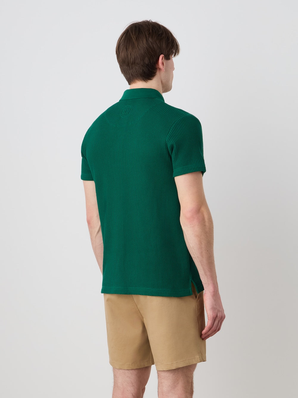 A man faces away from the camera, wearing the We Norwegians Waffle Polo Men – a relaxed fit green sweater – paired with tan shorts, set against a plain light-colored background.