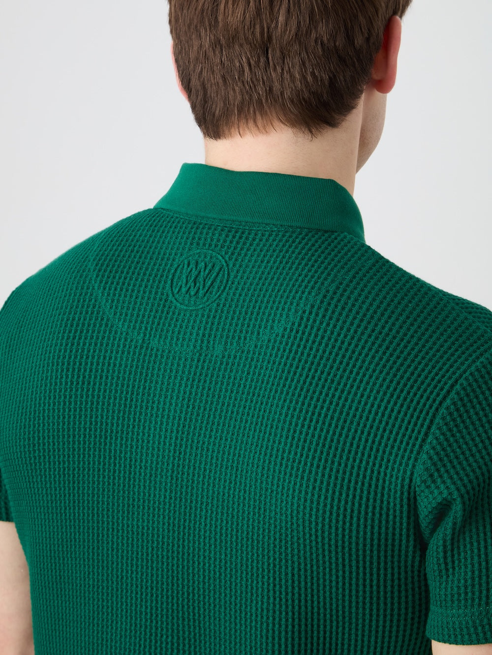 A person with short brown hair is seen from behind wearing the We Norwegians Waffle Polo Men, a textured green relaxed fit sweater featuring a small embroidered circular logo below the collar on the upper back.
