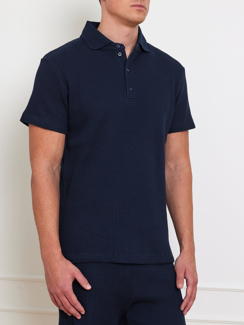 A man wearing We Norwegians' Waffle Polo Men, a textured waffle knit dark blue short-sleeve polo with button-up collar and relaxed fit, stands against a neutral background with his left hand casually by his side.