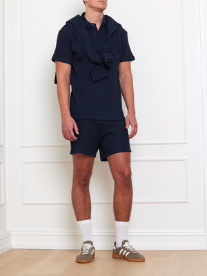 A man in a navy polo and sweater over his shoulders wears We Norwegians Waffle shorts in navy, white socks, and brown sneakers while standing against a white paneled wall.