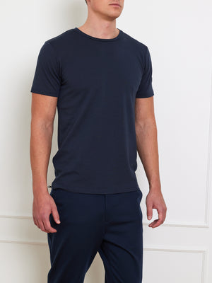 A person in a plain, dark blue Medalist Piqué Tee Men by We Norwegians and matching pants stands against a neutral, light-colored wall. The short-sleeved shirt exudes casual minimalism, and the piqué merino wool adds a subtle touch of sophistication. The person’s face remains unseen.
