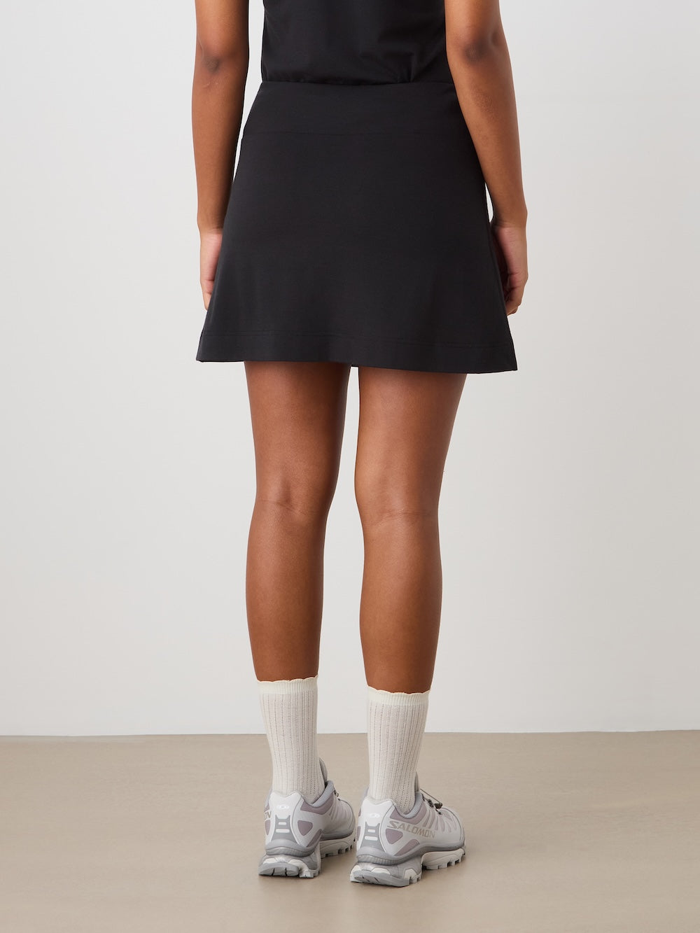 A person stands facing away, wearing the We Norwegians Medalist Piqué Skirt for women in black, paired with a black top, white socks, and gray sneakers against a plain light-colored background.
