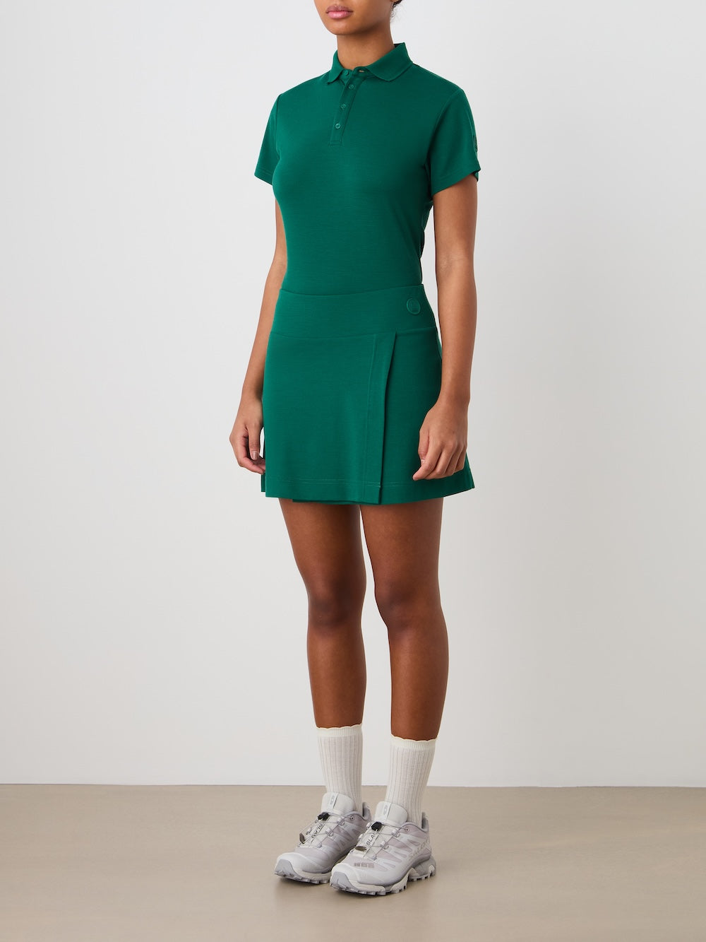 A person stands against a plain background wearing the We Norwegians Medalist Piqué Skirt Women, paired with a green short-sleeve polo, white socks, and gray athletic shoes. Their hands are relaxed by their sides.
