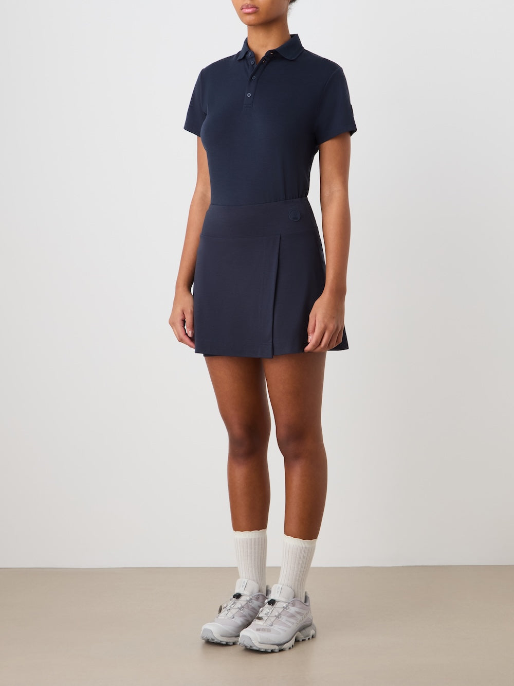 A person stands against a plain background wearing a navy We Norwegians Medalist Piqué Skirt and polo, with white socks and gray sneakers. Their arms are by their sides and their face is partially out of view.