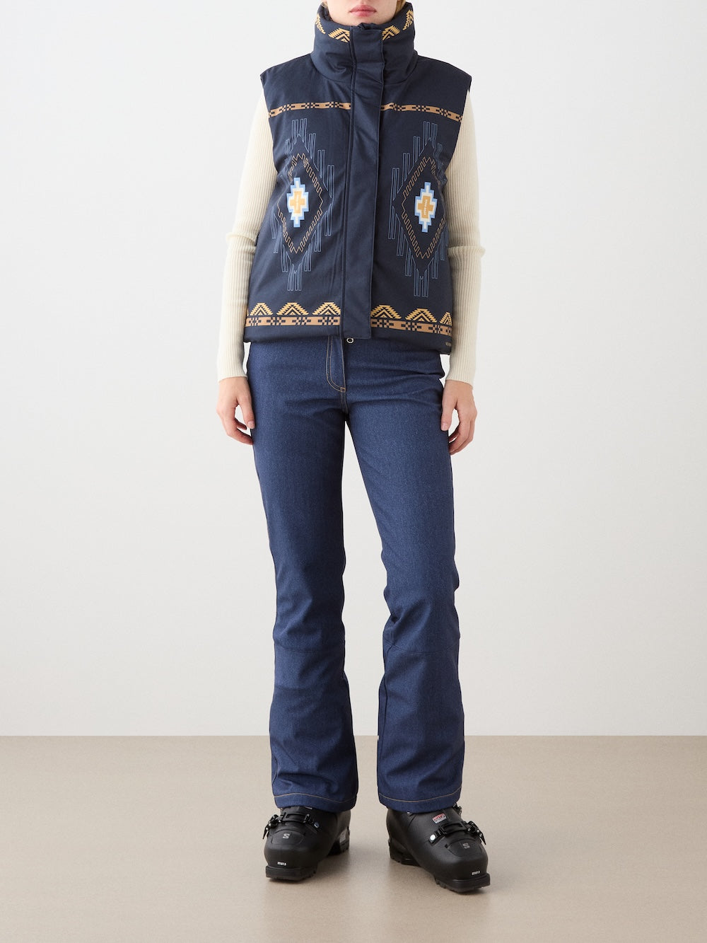 A woman wears the We Norwegians Pioneer Vest—a navy blue piece with blue, white, and gold geometric designs—over a cream turtleneck, jeans, and black winter boots. She stands indoors against a plain background.