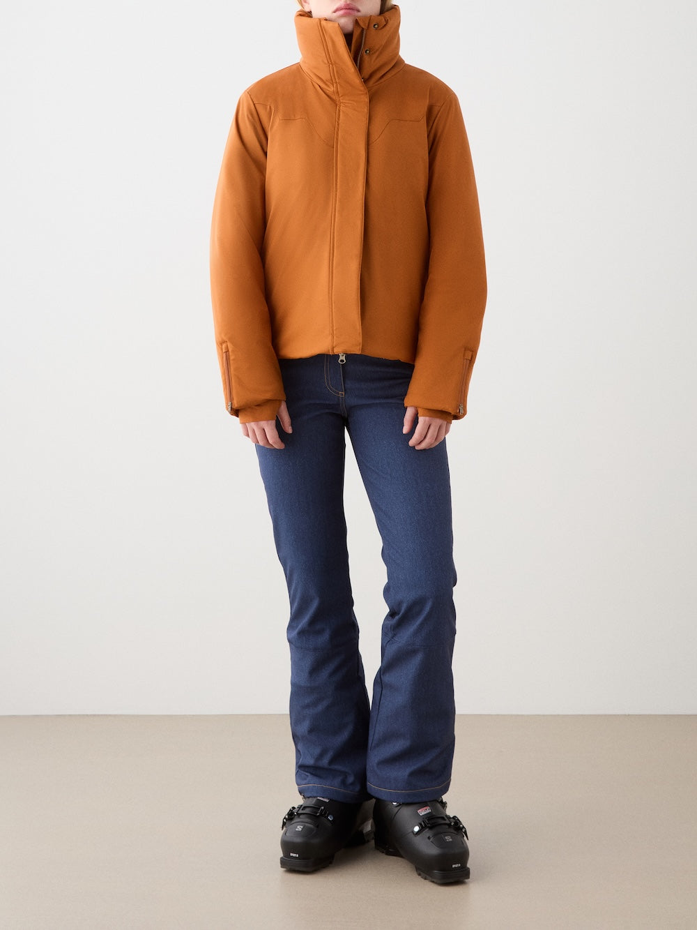 A person wears the We Norwegians Western Ski Jacket women with natural wool padding, blue jeans, and black ski boots, standing against a plain light backdrop with hands relaxed at their sides.