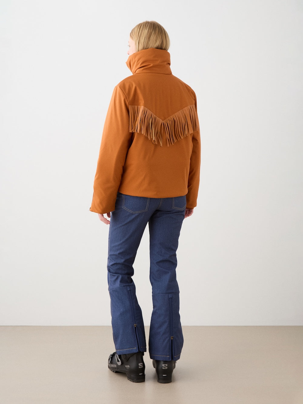 A person wears the We Norwegians Western Ski Jacket women with fringes across the back, paired with blue jeans and black boots. The high-collared jacket features natural wool padding for an alpine-inspired indoor look.