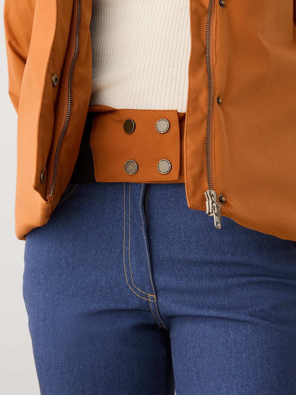 A person wears the We Norwegians Western Ski Jacket women in brown with natural wool padding, over a white ribbed shirt and blue jeans with contrast stitching. The unzipped jacket reveals a wide belt detail with four metallic snaps at the waist.