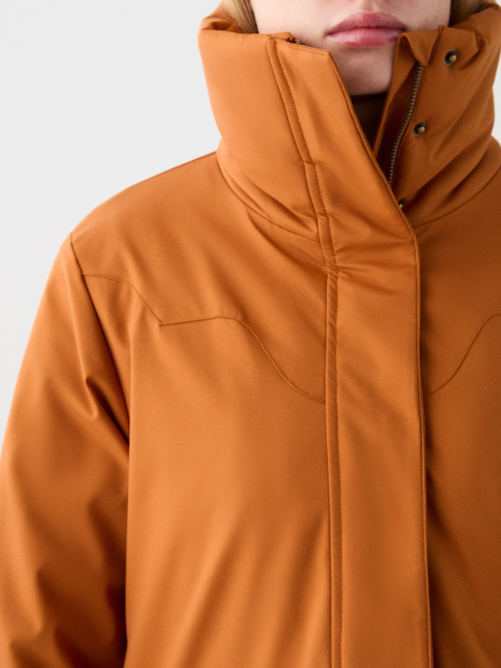 A person wears the We Norwegians Western Ski Jacket women in orange, with a high collar partially covering their mouth and neck. The jacket features subtle stitching, snap buttons, and natural wool padding; only the upper torso and lower face are visible.