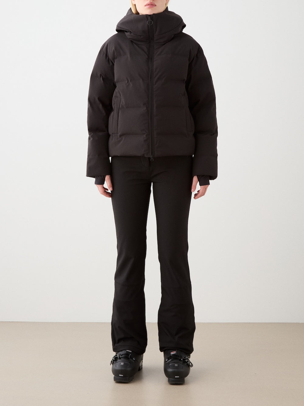 A person stands facing forward in a We Norwegians Aurora Ski Jacket for women, paired with black fitted pants and black ski boots, against a plain light background. Their face is partially out of frame.
