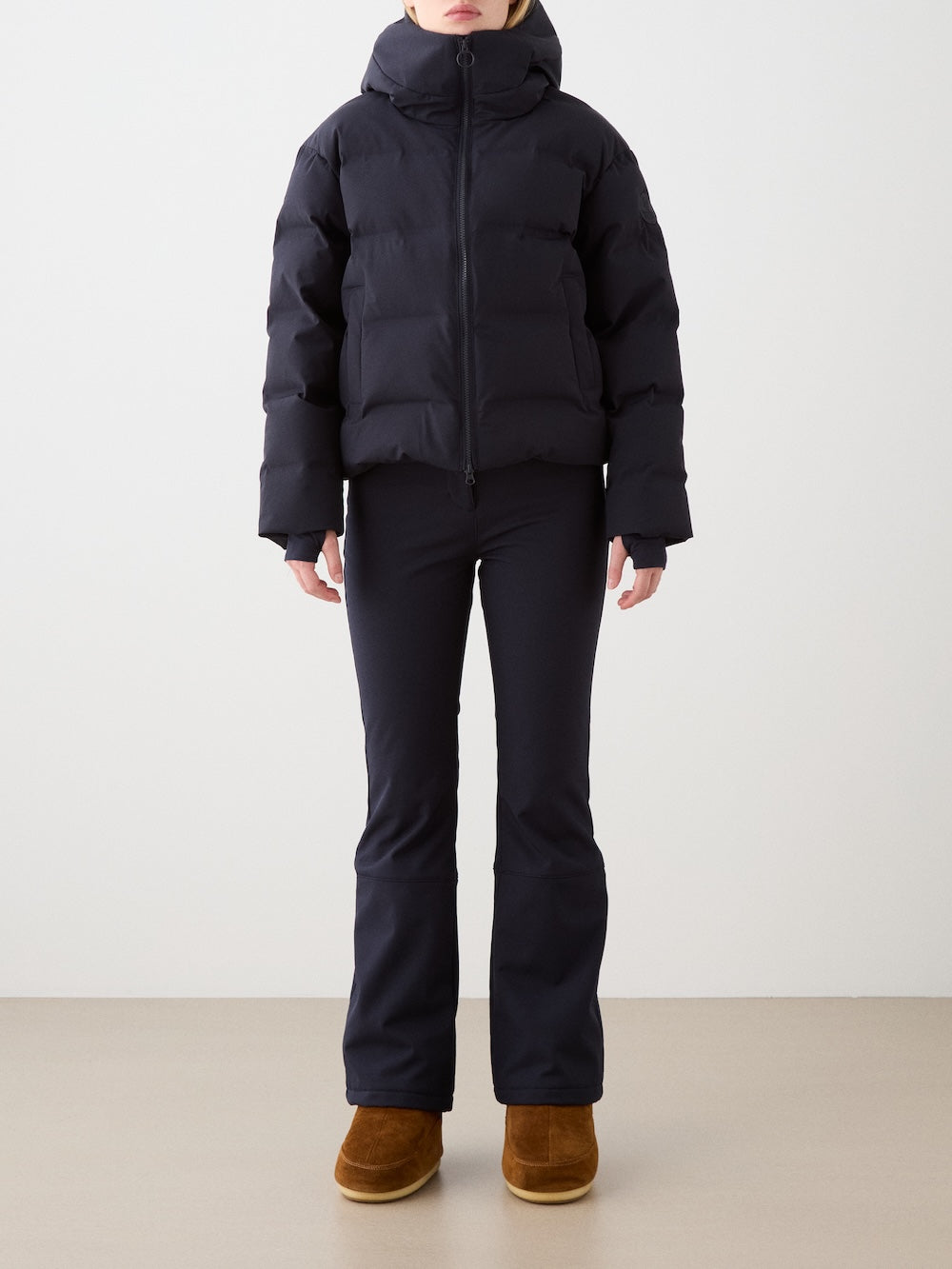 A model in a black We Norwegians Aurora Ski Jacket for women, black flared pants, and brown suede boots stands against a plain white background, with their face not visible.