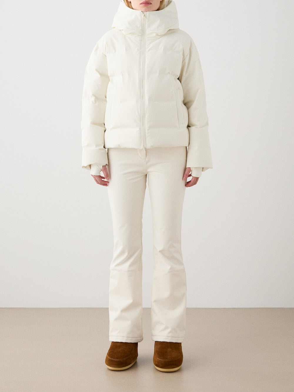 Dressed in the We Norwegians Aurora Ski Jacket women in off-white with a hood, flared pants, and brown boots, she stands against a plain, light background. The responsible down, waterproof jacket offers both warmth and style.