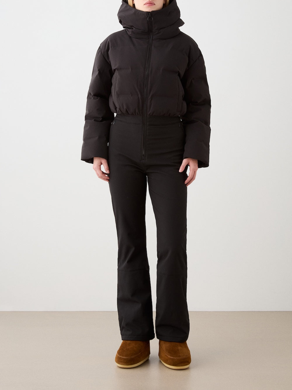 A woman stands facing forward against a plain light background, wearing the We Norwegians Aurora Ski Suit in black, with arms relaxed at her sides.