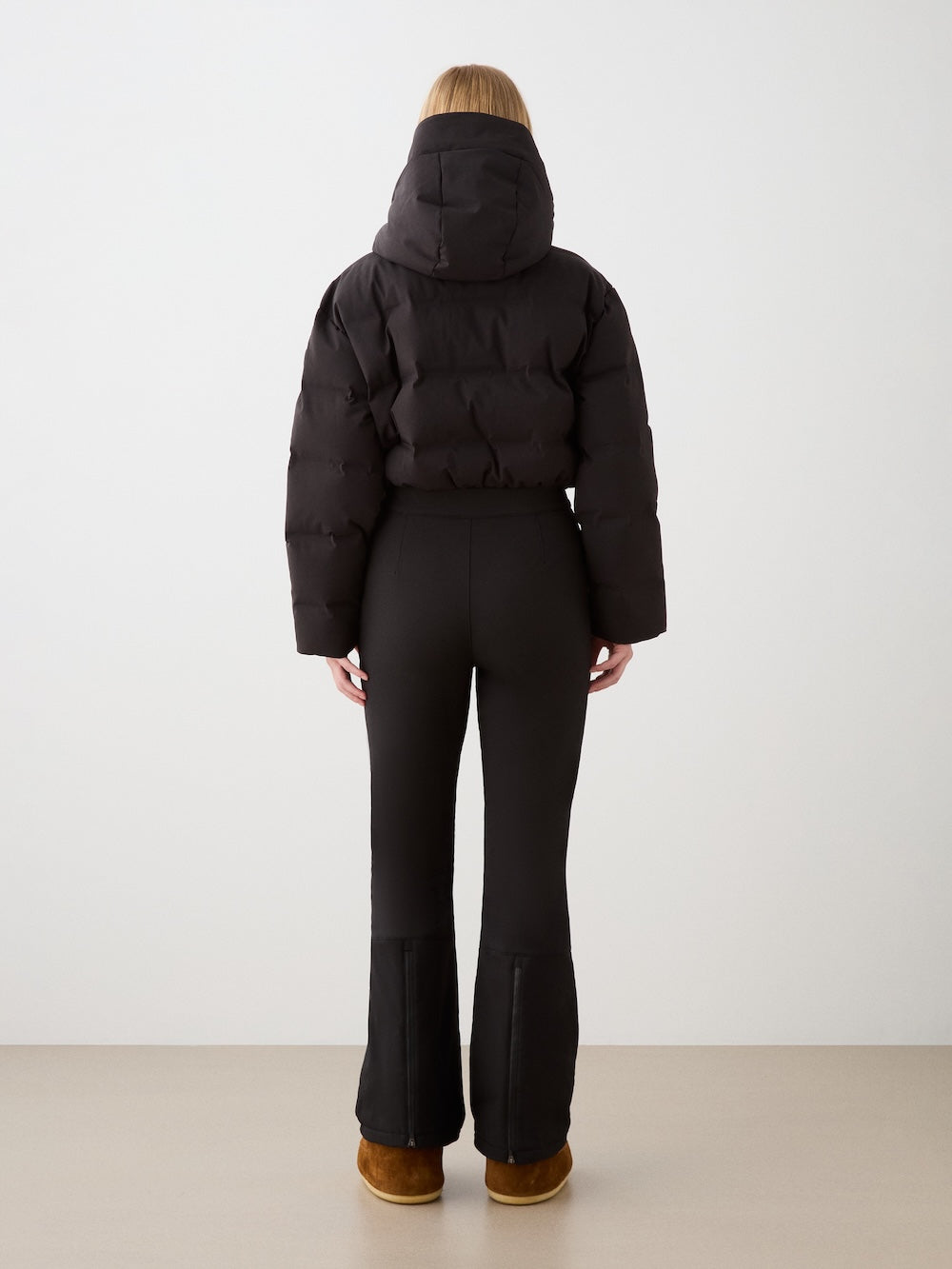 A person with long blonde hair wears the We Norwegians Aurora Ski Suit for women, standing with their back to the camera against a light backdrop, showcasing a sleek alpine style.