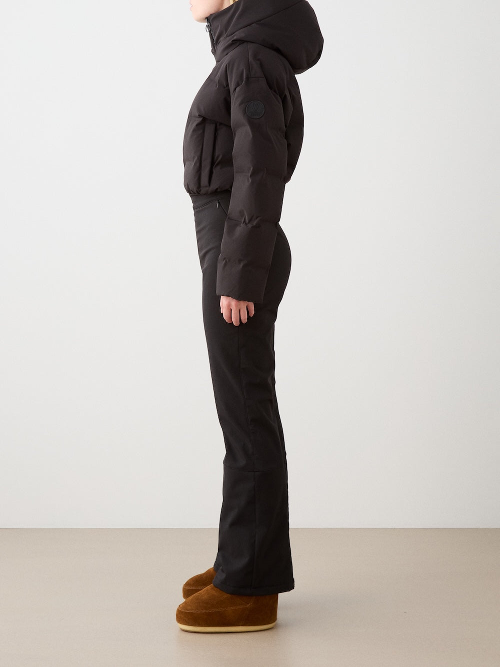 A woman stands in profile against a plain background, wearing the We Norwegians Aurora Ski Suit for a sleek alpine look.