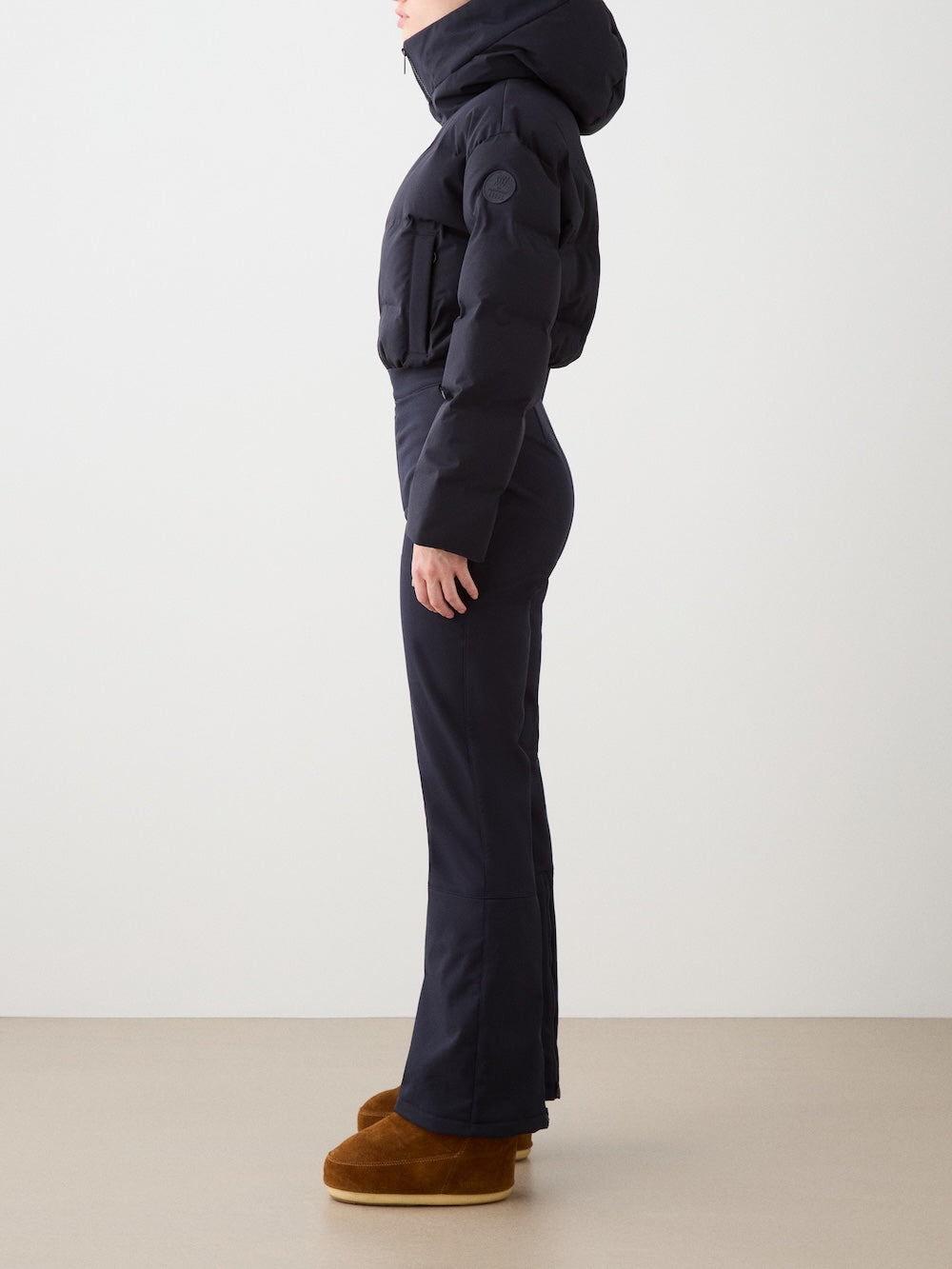 A person stands in profile against a plain light background, wearing the We Norwegians Aurora Ski Suit for women—a black puffer jacket with a hood, paired with fitted dark pants and brown platform boots.