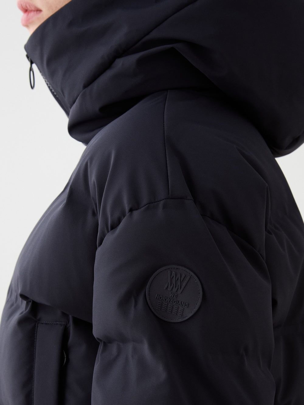 Close-up of a person wearing the We Norwegians Aurora Ski Suit for women. The dark suit features a hood and a circular patch with text and emblem on the upper sleeve, against a plain light background. Made with responsible down.