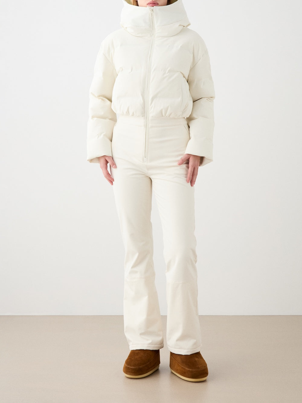 Wearing the We Norwegians Aurora Ski Suit, a woman stands against a plain light background. Her face is partly out of frame, highlighting the chic alpine style of this all-white ski suit ensemble.