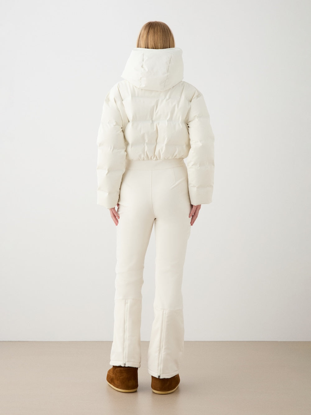 A woman with straight blonde hair, seen from behind in We Norwegians’ Aurora Ski Suit, off-white and alpine-inspired, paired with brown boots, stands against a plain white wall.
