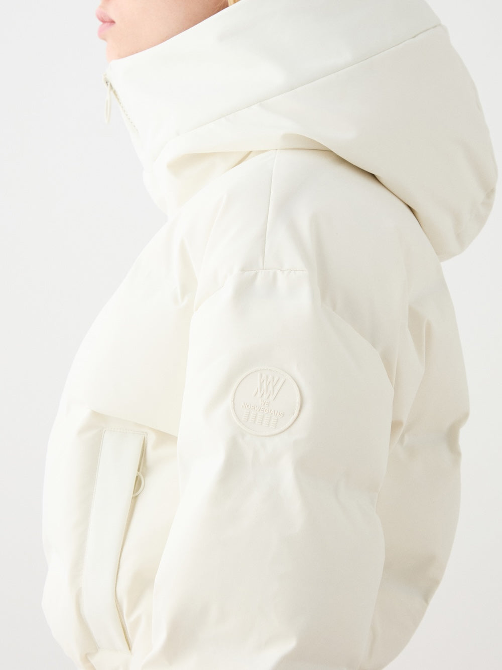 A person wears the We Norwegians Aurora Ski Suit for women—a white, hooded puffer with a circular sleeve patch, responsible down filling, and a flap front pocket. The alpine-inspired look is shown with the wearer’s face partially out of frame.