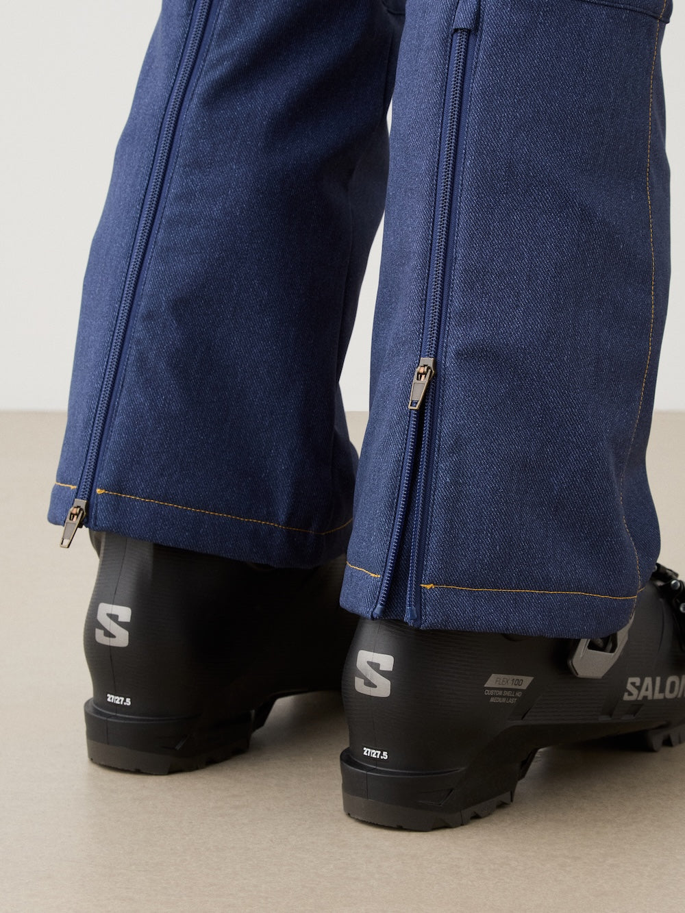 Close-up of a person wearing We Norwegians Western Ski Pant women in blue, featuring cuff zippers partially open over black Salomon ski boots. Photo shows lower legs and feet from behind.