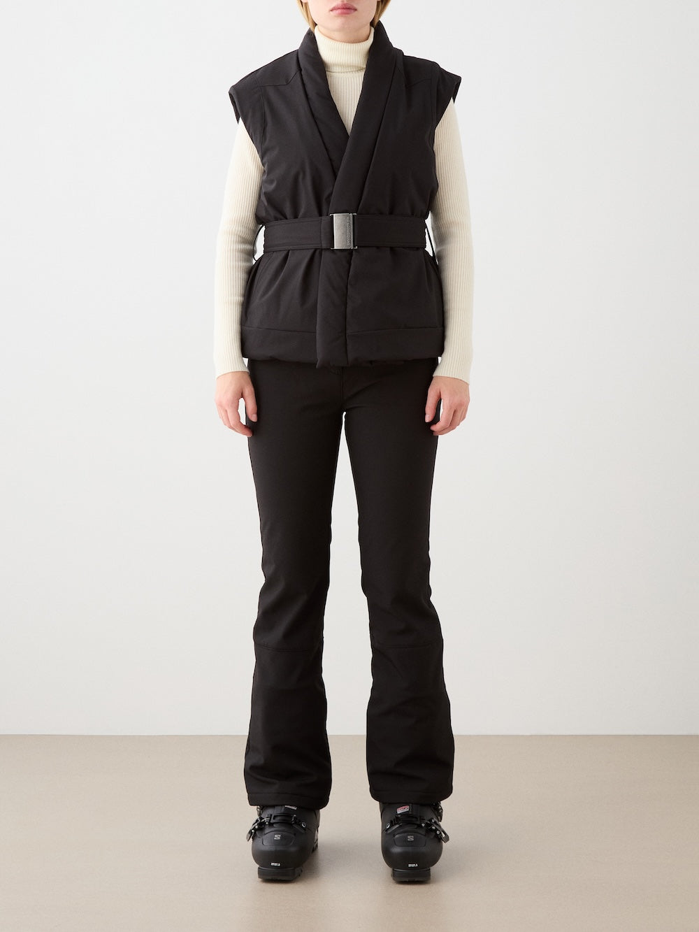 A person faces forward in a We Norwegians Western Vest for women—a belted black wool padded vest—layered over a cream turtleneck, paired with black pants and ski boots. Their face is not visible, set against a plain light background.