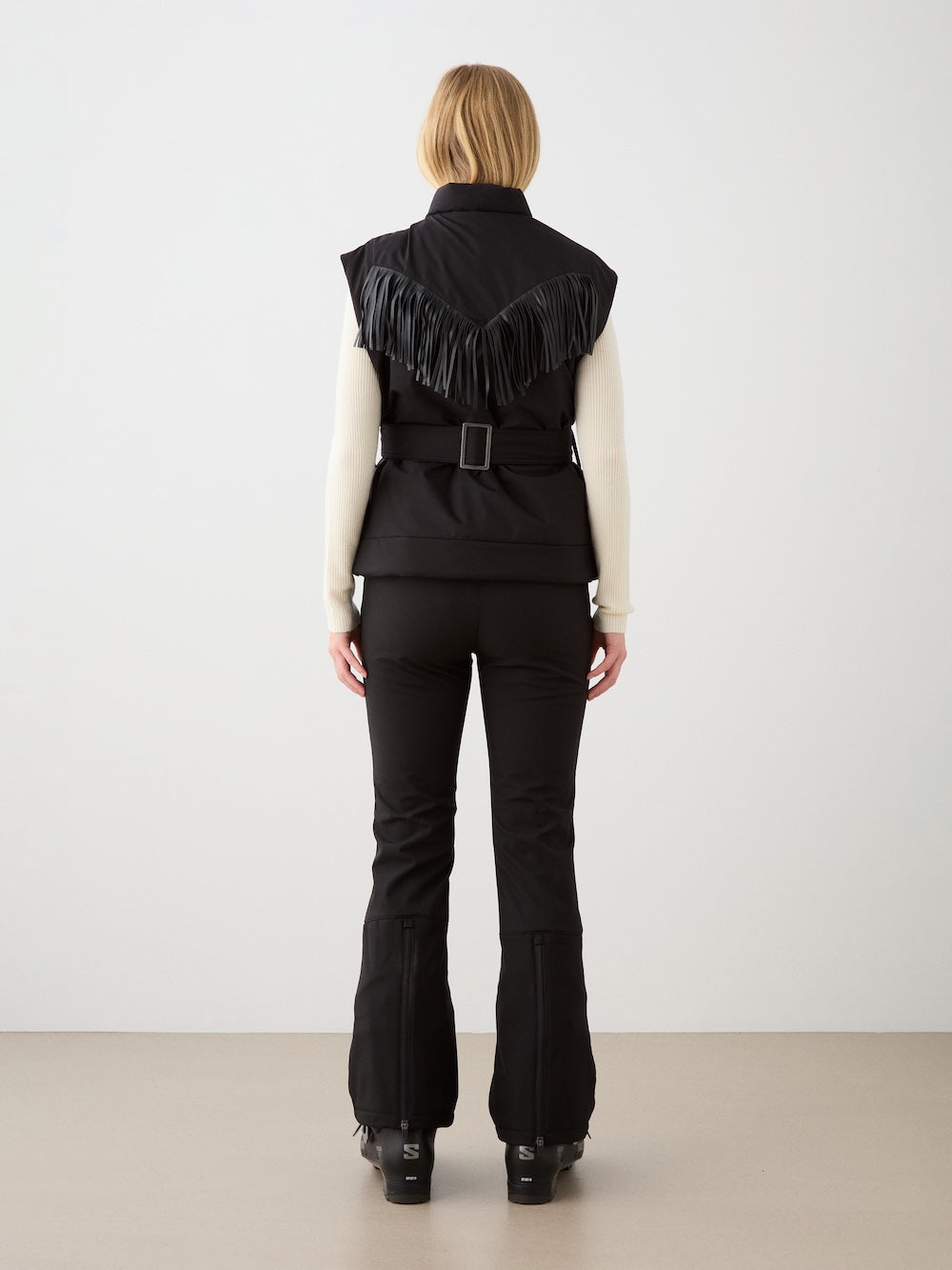 A woman with short blonde hair faces away, wearing the We Norwegians Western Vest for women—black, wool, padded with fringe detail—styled with a white long-sleeve top, black belted pants, and black shoes against a plain indoor backdrop.