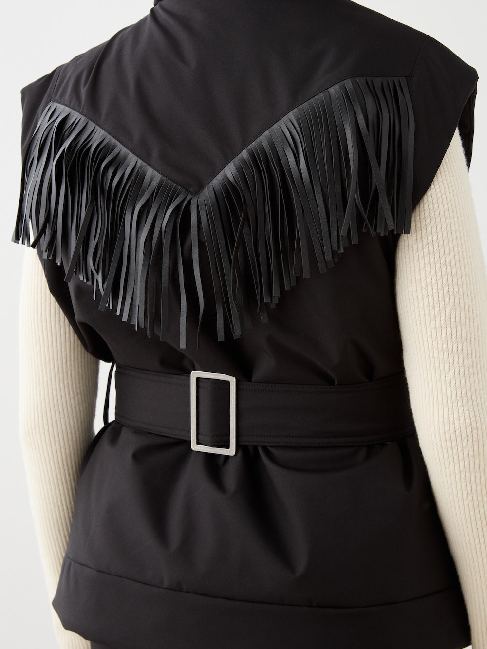 Close-up of a person wearing the We Norwegians Western Vest for women, featuring long black fringe on the back and a wide belt with silver buckle, layered over a cream ribbed long-sleeve top.