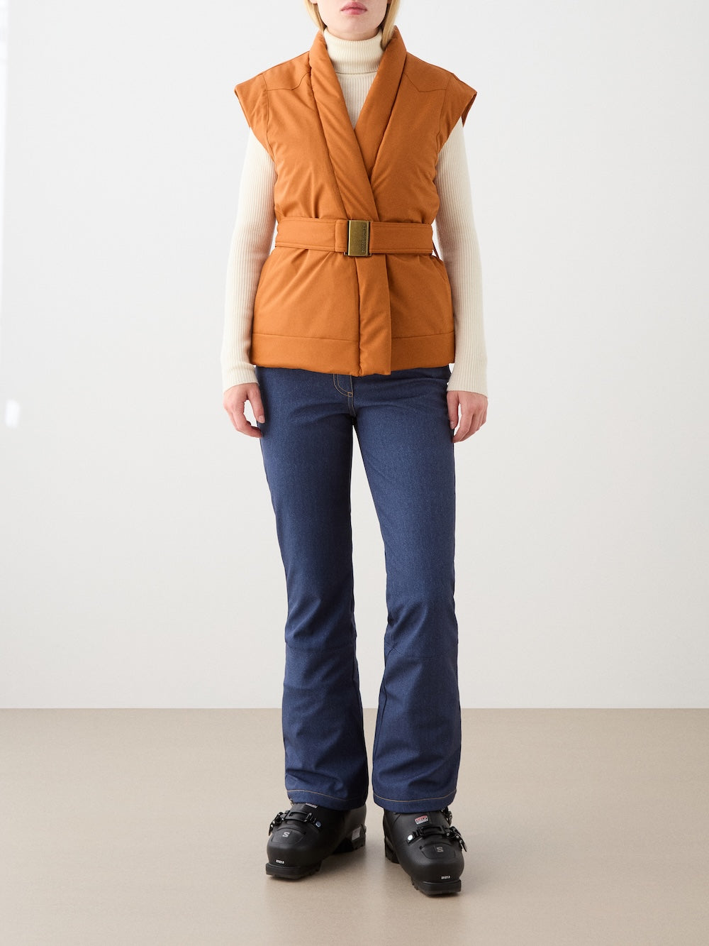 A woman stands indoors wearing the We Norwegians Western Vest over a white turtleneck, blue jeans, and black ski boots against a minimalistic background.