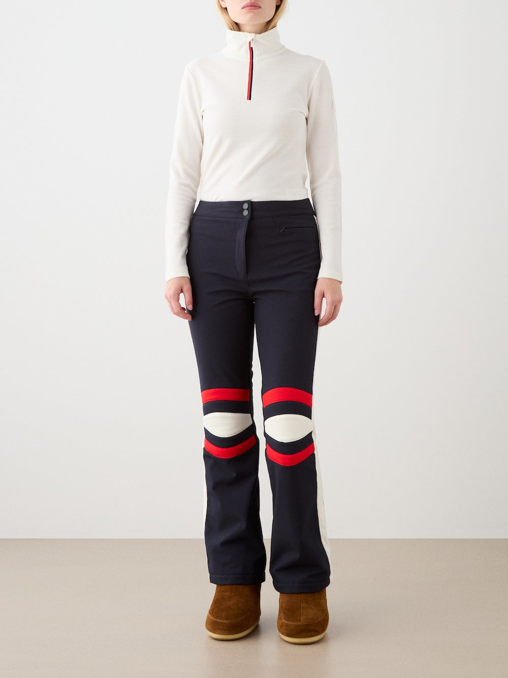A person stands against a plain background wearing a white long-sleeve turtleneck, brown boots, and We Norwegians Solo Ski Pant for women with padded knees and red-and-white circular designs. The person's face is not visible.