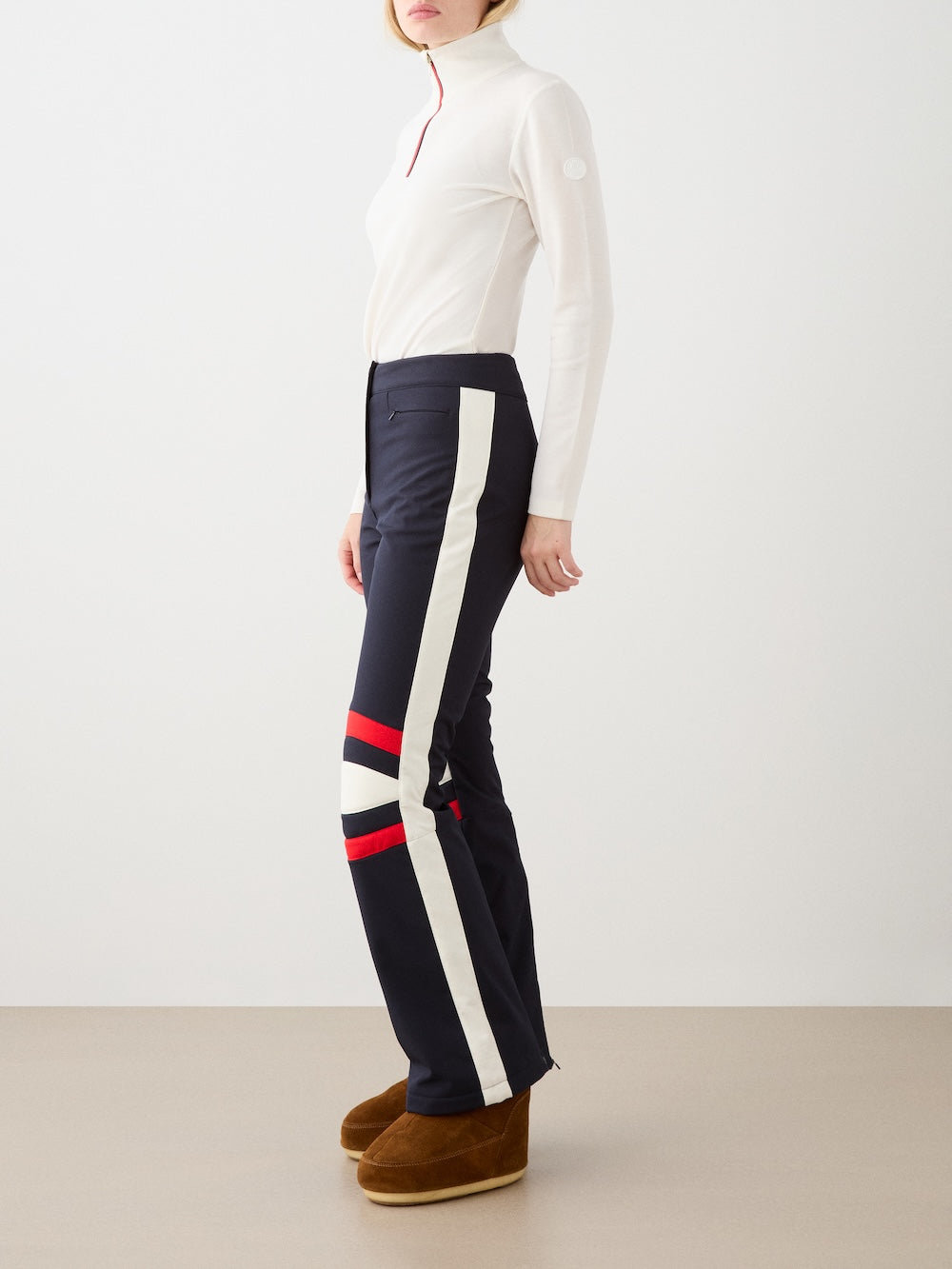 A woman in profile wears the We Norwegians Solo Ski Pant for women—featuring white and red stripes—paired with a white turtleneck and brown platform boots, posed against a plain backdrop.