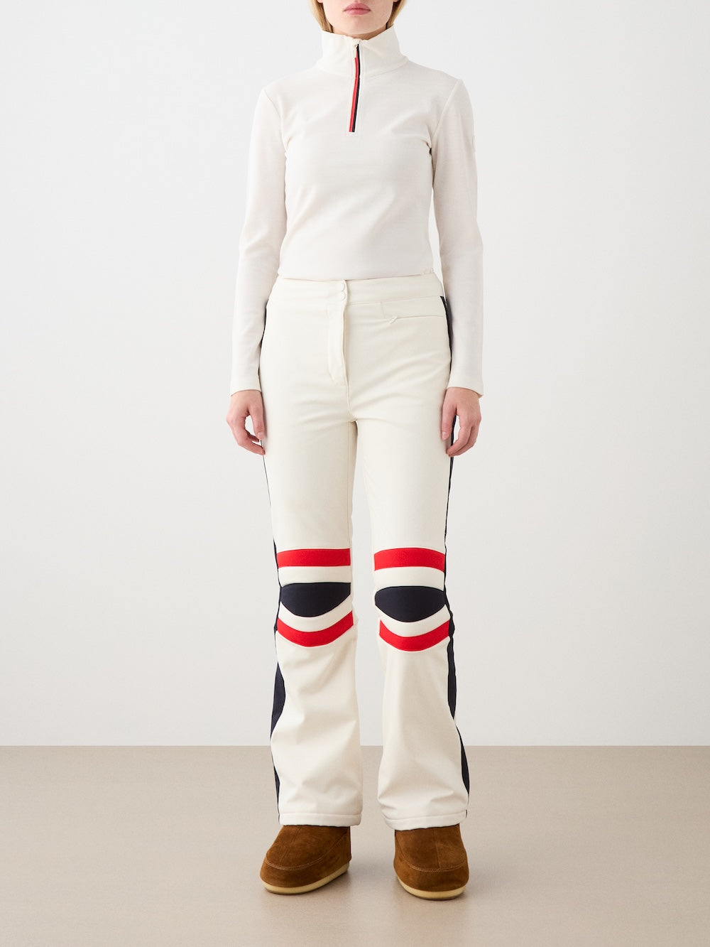 A person stands against a plain background wearing We Norwegians Solo Ski Pant for women, featuring a cream color with retro red and black stripes and padded knees, paired with a white zip turtleneck and brown boots. Their face is not visible.
