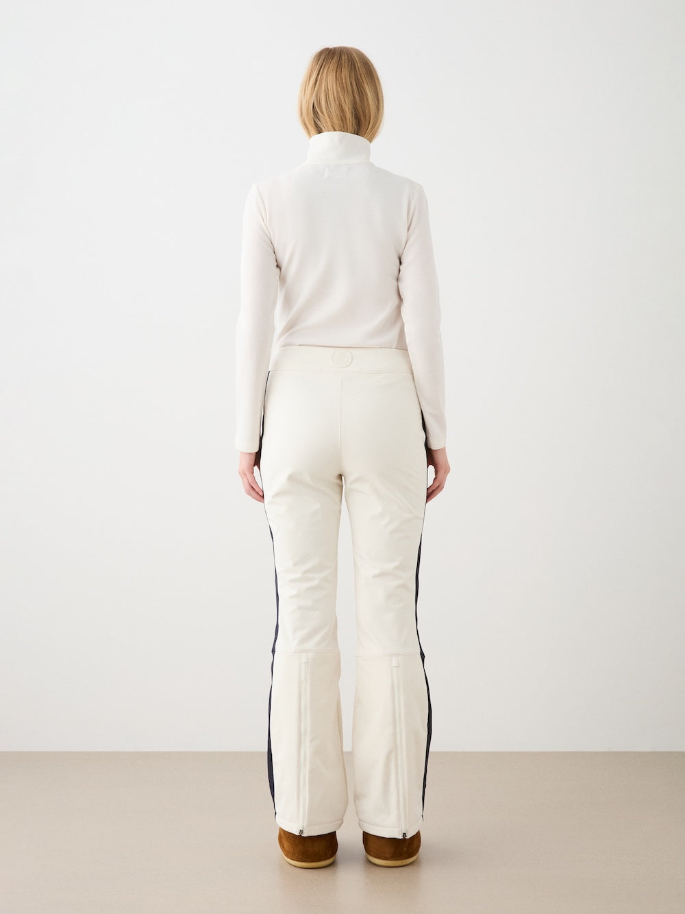 A blonde woman in a white turtleneck and We Norwegians Solo Ski Pant women—cream with black side stripes and padded knees—stands facing away from the camera against a plain background, wearing brown shoes.