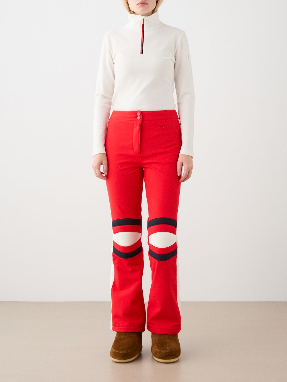 A person wears We Norwegians’ Solo Ski Pant for women—featuring bright red color, circular knee cutouts, and black details—with a white zip-up top and brown boots, standing against a plain light backdrop.