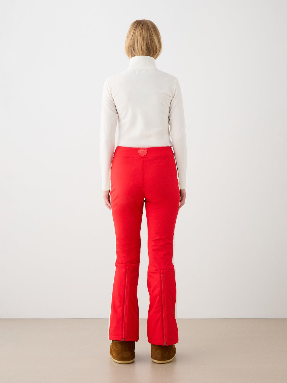A woman faces away, wearing We Norwegians Solo Ski Pant in bright red with padded knees, a white turtleneck, and brown boots against a plain light background.
